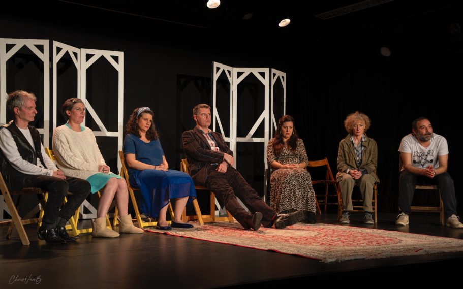 Huit personnes assises sur des chaises, sur scène avec un décor minimaliste.